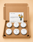 An open kraft cardboard gift box containing six small white wax candles with round labels and a white insert card reading 'Flora + Moon Autumn Collection 2020' with a floral illustration, set on a warm beige background.