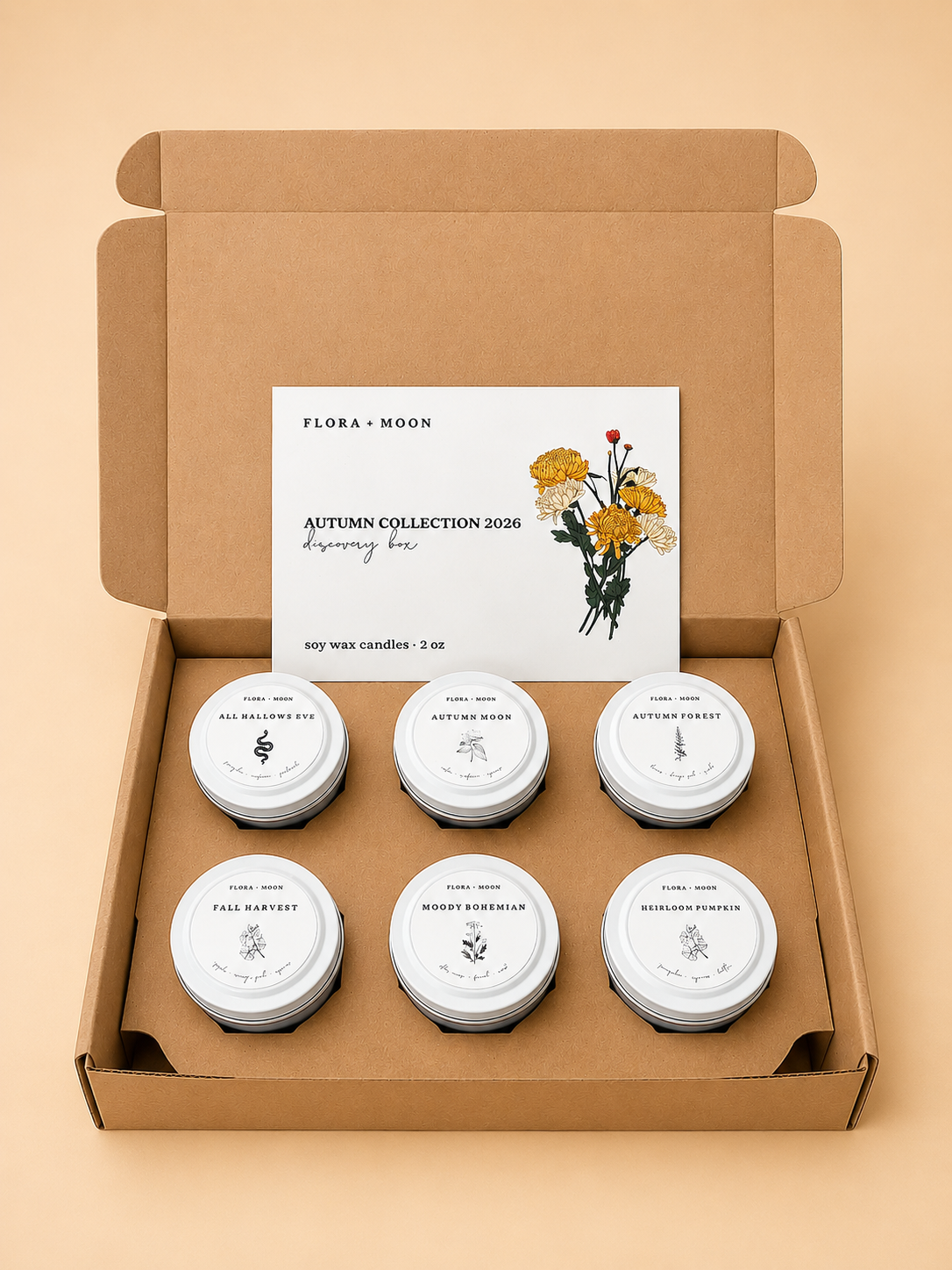 An open kraft cardboard gift box containing six small white wax candles with round labels and a white insert card reading 'Flora + Moon Autumn Collection 2020' with a floral illustration, set on a warm beige background.