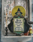 Witch-themed decor with a framed picture of a witch holding a sign labeled 'Witch Services' on a shelf.